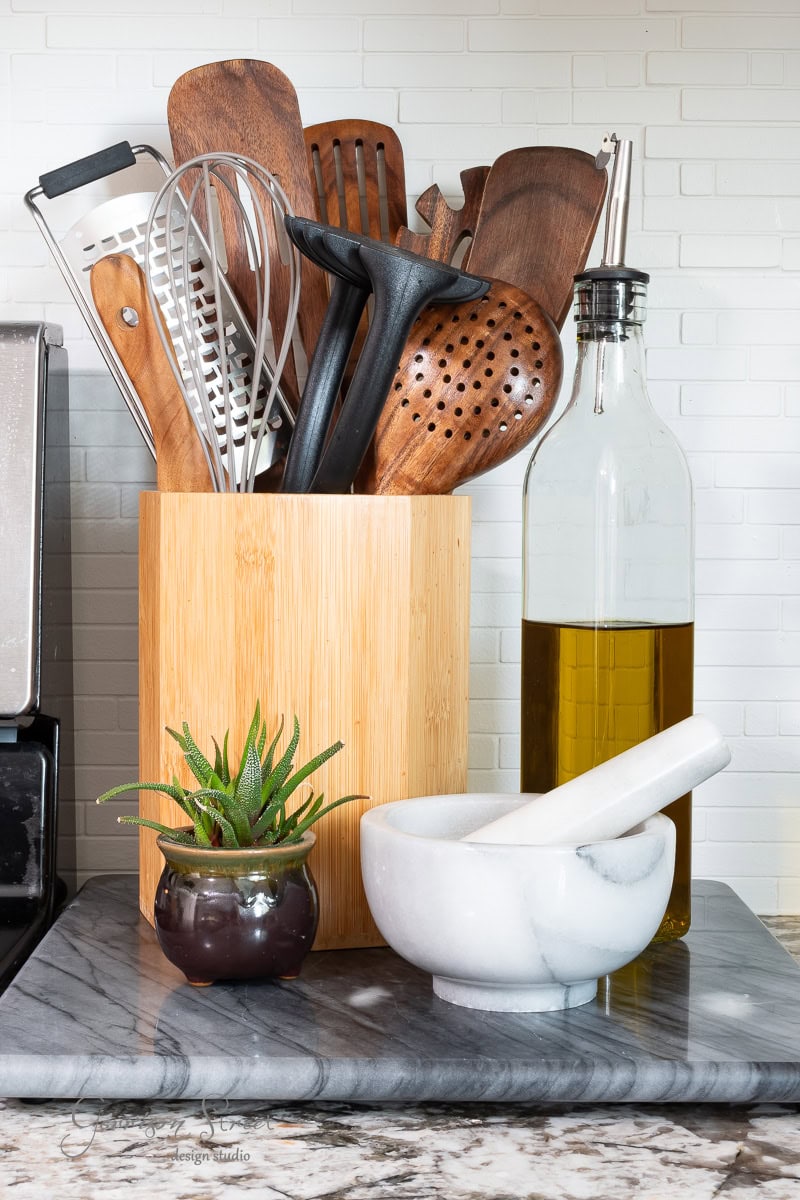 A kitchen countertop features a bamboo holder with wooden utensils, a neatly displayed cutting board, a bottle of olive oil, a small potted succulent, and a white marble mortar and pestle. The scene is completed by a white tiled backsplash that adds brightness to the space.