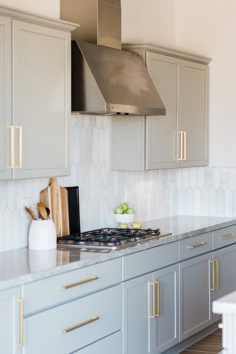 A modern kitchen showcases light gray cabinets with gold handles, a stainless steel range hood, and a gas stove. A lesson in styling, the marble countertop features cutting boards elegantly displayed alongside a vase and a bowl of green apples against the geometric tile backsplash.