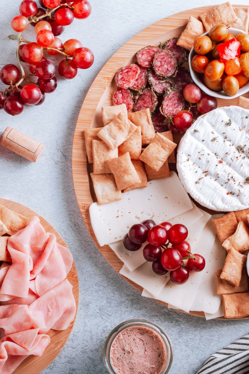 A charcuterie board with brie cheese, salami, olives, grapes, and crackers illustrates how to display cutting boards on kitchen counters. Nearby, sliced ham is placed on another plate. A glass jar with pâté and a bunch of red grapes complete the scene on a light textured surface.