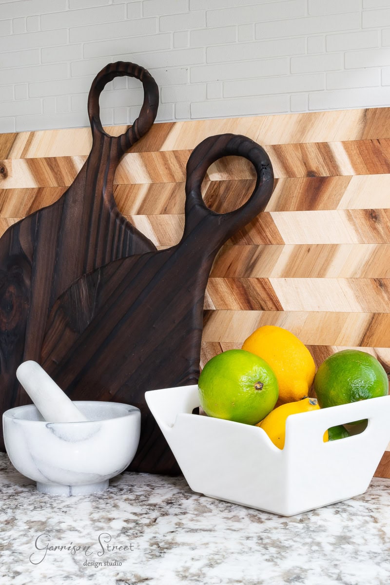 A kitchen counter showcases how to display cutting boards with a herringbone wooden backsplash, featuring two large boards in contrasting wood tones. In front are a white mortar and pestle and a white ceramic bowl filled with lemons and limes, adding vibrant color to the countertop ensemble.