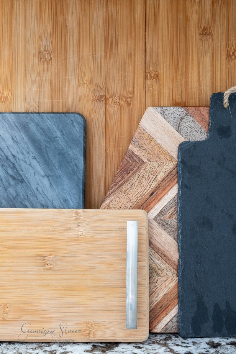 Various cutting boards, artfully displayed against a wooden wall, show off diverse materials like slate, bamboo, and wood with a herringbone pattern. For inspiration on how to display cutting boards on kitchen counters, observe the granite countertop beautifully tying it all together.