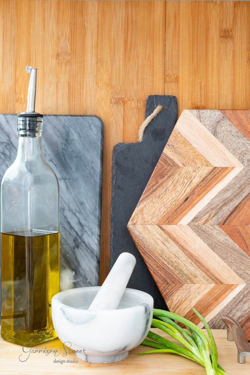 A kitchen scene beautifully illustrates how to display cutting boards on kitchen counters, showcasing a bottle of olive oil, a marble mortar and pestle, several cutting boards in varying wood tones, and an aloe plant on a wooden countertop.