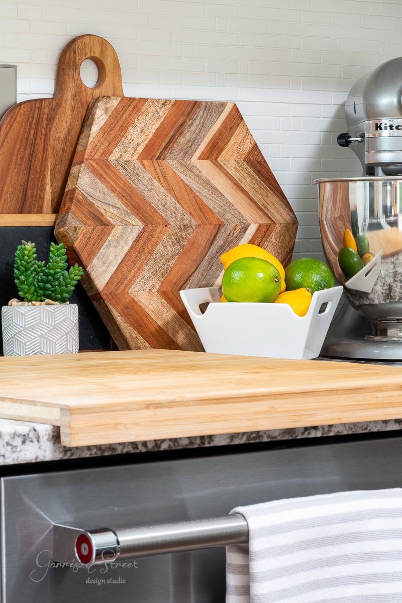 A modern kitchen counter showcases how to display cutting boards artfully, featuring a wooden cutting board and a chevron-patterned one. A white bowl of lemons and limes, a potted succulent, and a stand mixer grace the granite surface with a subtle glimpse of the stovetop oven.