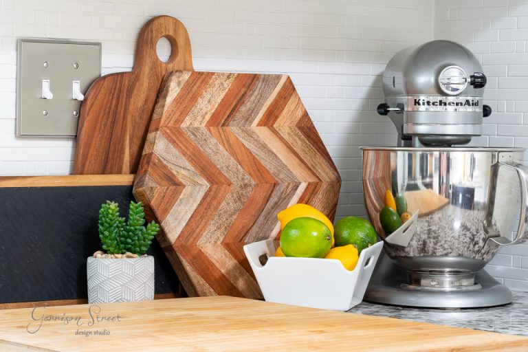 A kitchen counter showcases a mixer alongside wooden cutting boards, artfully arranged for those curious about how to display cutting boards on kitchen counters. A decorative plant and a vibrant bowl of lemons and limes add color against the backdrop of a white tile backsplash and light switch plate.