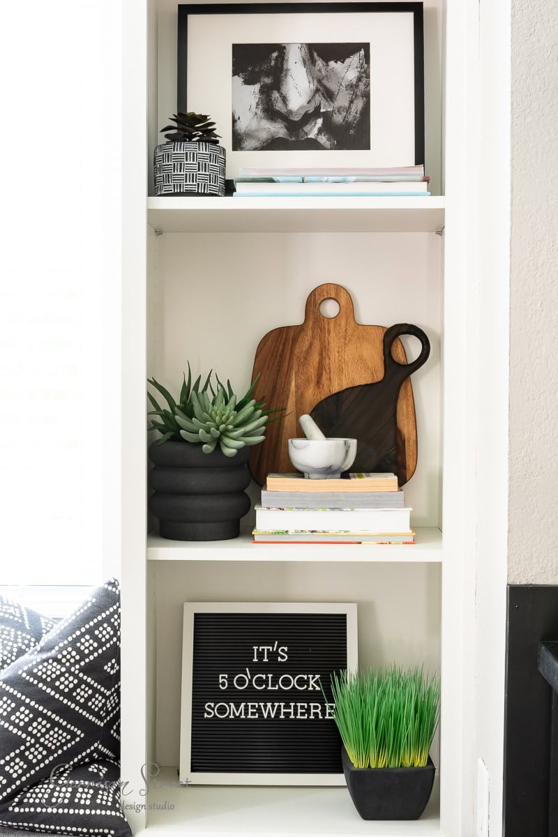 A shelf with framed art, decorative plants, and a cutting board perfectly illustrates how to display cutting boards on kitchen counters. Books and stacked bowls sit below, enhancing the cozy vibe. A sign reads, "It's 5 o'clock somewhere," while a patterned cushion peeks out on the left.