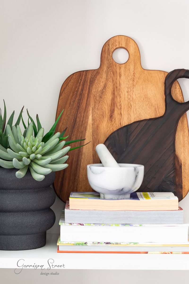 A wooden cutting board, a marble mortar and pestle, and a black sculptural planter with a green succulent illustrate how to display cutting boards on kitchen counters. Decorative books are stacked nearby, completing the elegant shelf arrangement.