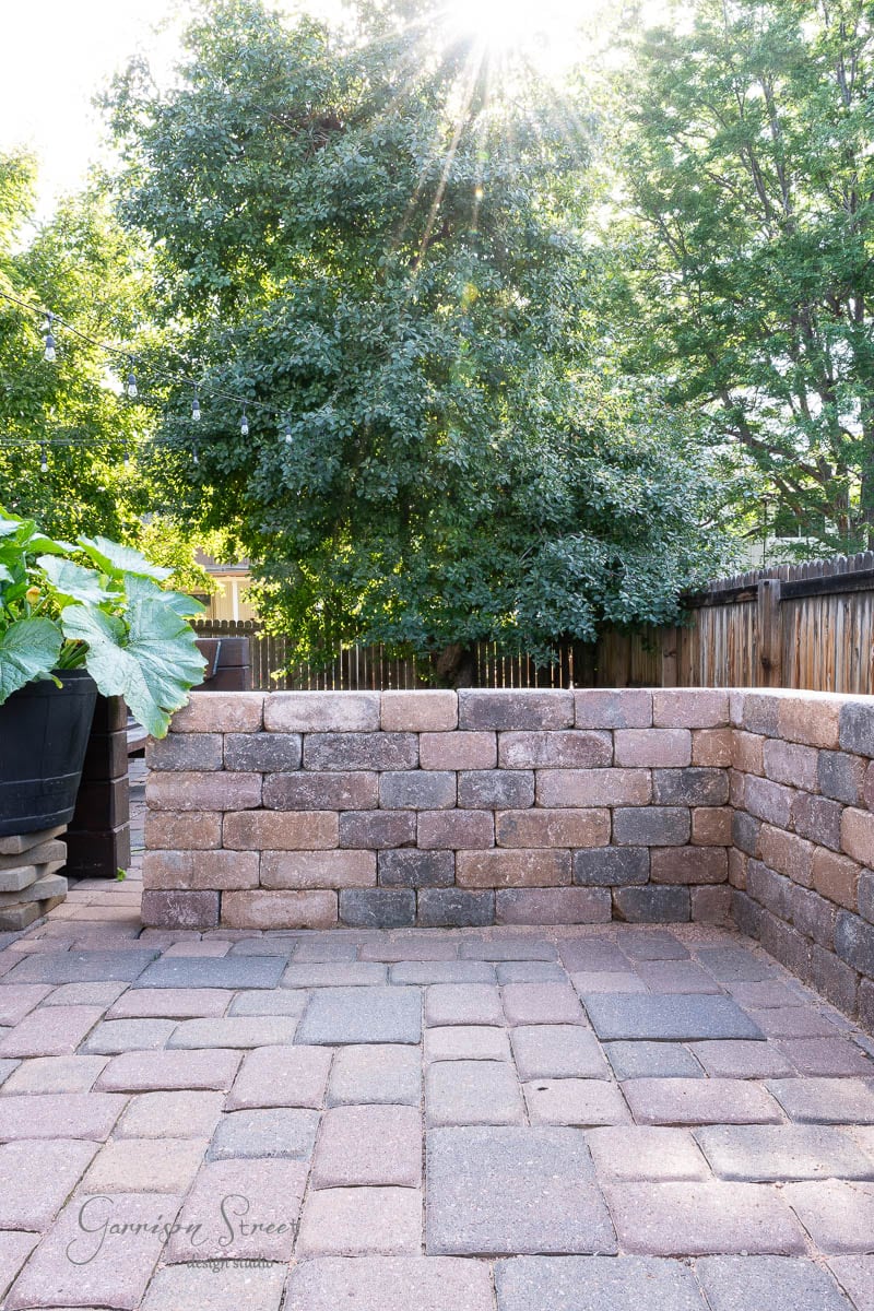 A sunlit backyard scene showcases a stone patio bordered by short brick walls. Within this inviting space, a DIY Traeger outdoor kitchen takes center stage. Lush green trees and plants surround the area, with sunlight streaming through the leaves, while a wooden fence encloses it all.