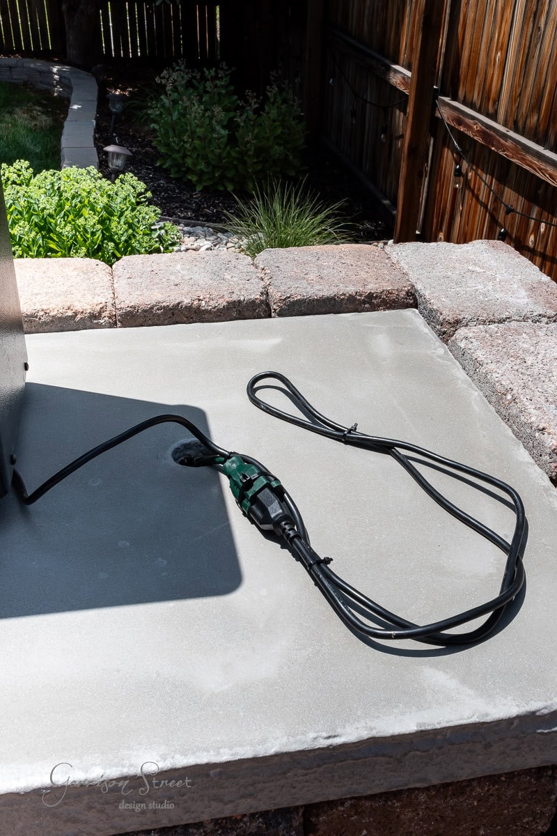A coiled black electrical cord with a green connector rests on a smooth concrete surface, hinting at a DIY project in progress. The background features a garden with shrubs, a wooden fence, and the perfect spot for your Traeger outdoor kitchen setup.