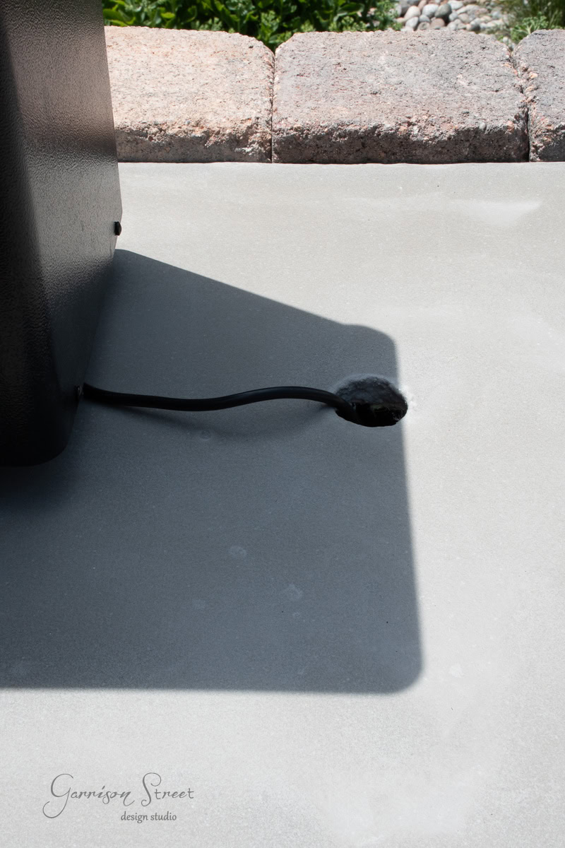 A black cable extends from a dark metallic box into a small hole in light gray concrete, adding a sleek touch to your DIY Traeger outdoor kitchen. Sunlight casts a shadow of the box on the concrete surface. In the background, a row of stone bricks borders green foliage.