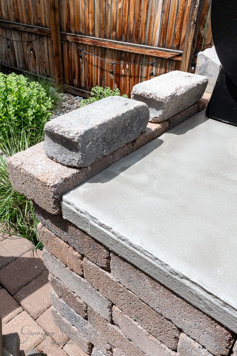 A close-up of a brick outdoor kitchen counter with a sleek concrete countertop showcases the craftsmanship of a DIY Traeger setup. Two gray bricks are placed on top. In the background, there's a wooden fence and lush green foliage, adding to the rustic charm.