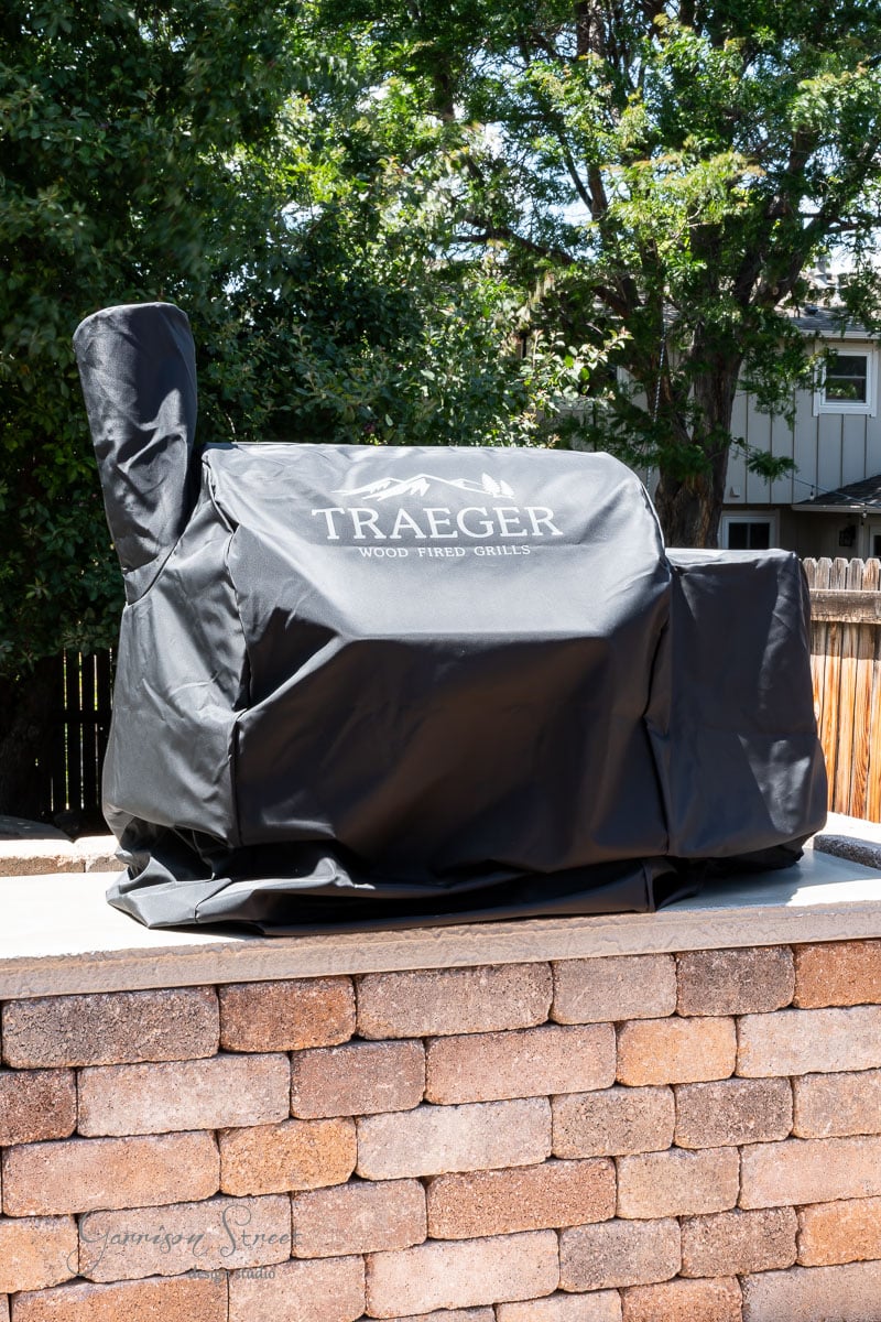 A covered Traeger wood-fired grill is the centerpiece of this DIY outdoor kitchen, sitting on a stone patio under the shade of trees. The black grill cover proudly displays the Traeger logo, with a wooden fence and house visible in the background.