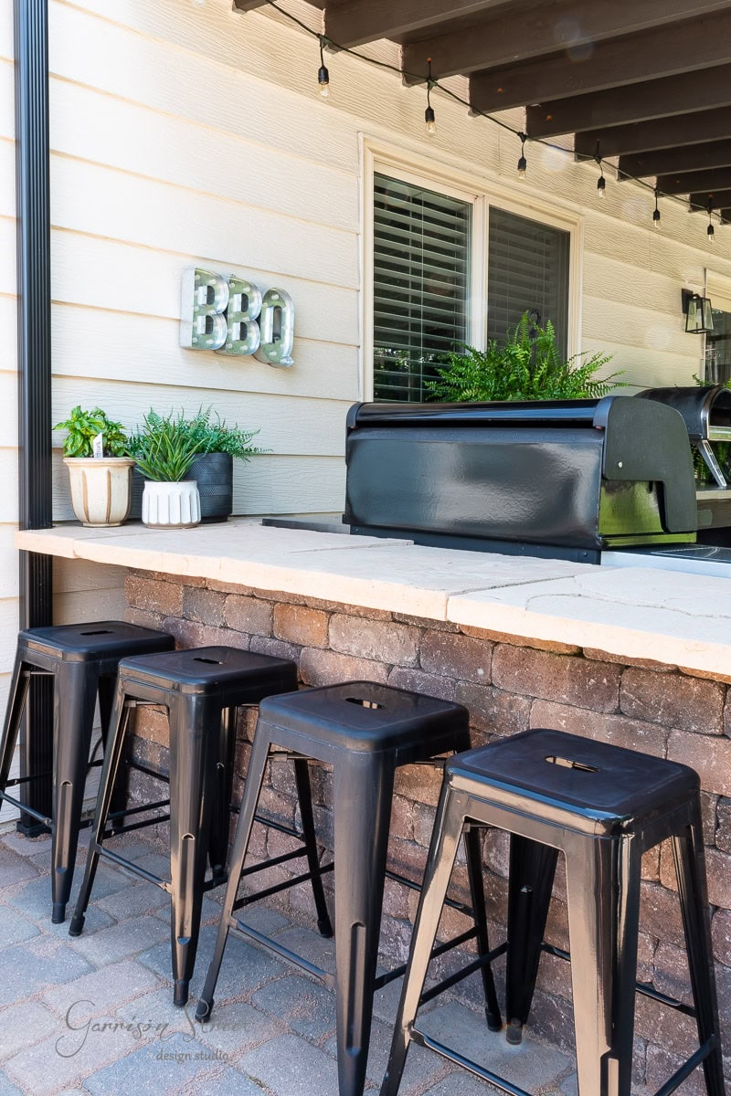 Outdoor patio area featuring a DIY Traeger grill setup, a counter with potted plants, and four black metal stools. String lights hang above, creating a cozy atmosphere. The siding of a house and a window are in the background.