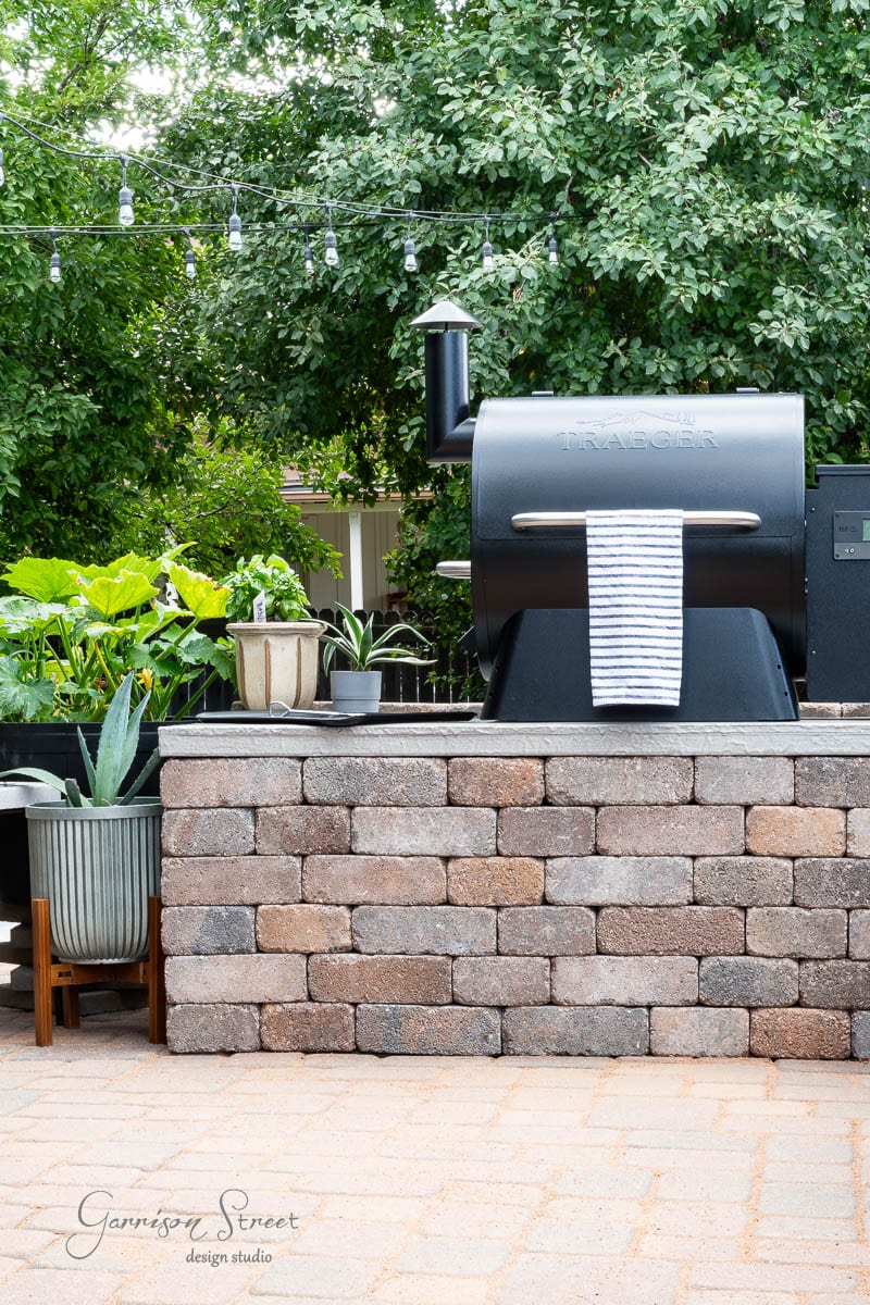 Outdoor patio scene with a black grill on a brick platform, part of a charming DIY outdoor kitchen. A striped towel hangs on the grill handle. Green plants in pots decorate the area, and string lights are strung overhead against a backdrop of lush trees.