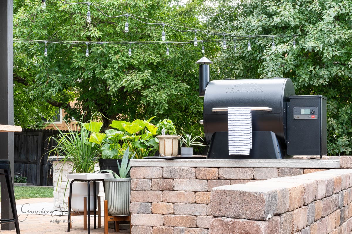 A DIY Traeger outdoor kitchen graces the backyard patio, where a robust stone counter hosts the grill. Potted plants and a white towel adorn it, while string lights twinkle above, framed by lush, towering trees.
