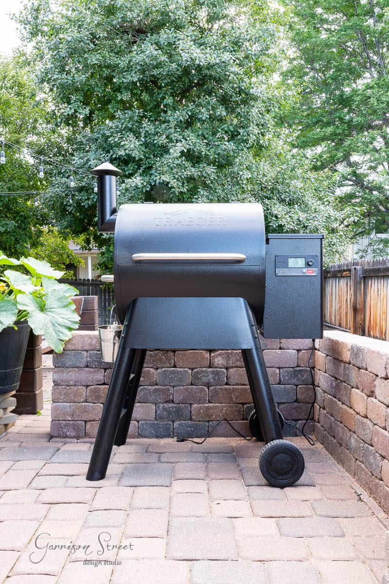 A black Traeger grill stands proudly on a stone patio, part of a DIY outdoor kitchen project. Large green plants are to the left, while a wooden fence frames the background. Trees provide shade, with sunlight filtering gently through the leaves.
