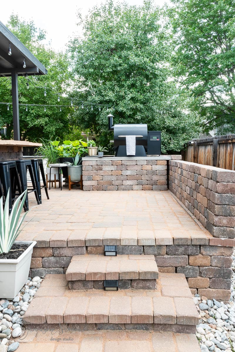 A patio features a DIY Traeger outdoor kitchen complete with a brick barbecue grill and bar with black stools. Steps lead down to a pebble walkway adorned with potted plants. Tall trees surround the area, creating a lush green backdrop, while string lights hang above, adding a cozy atmosphere.