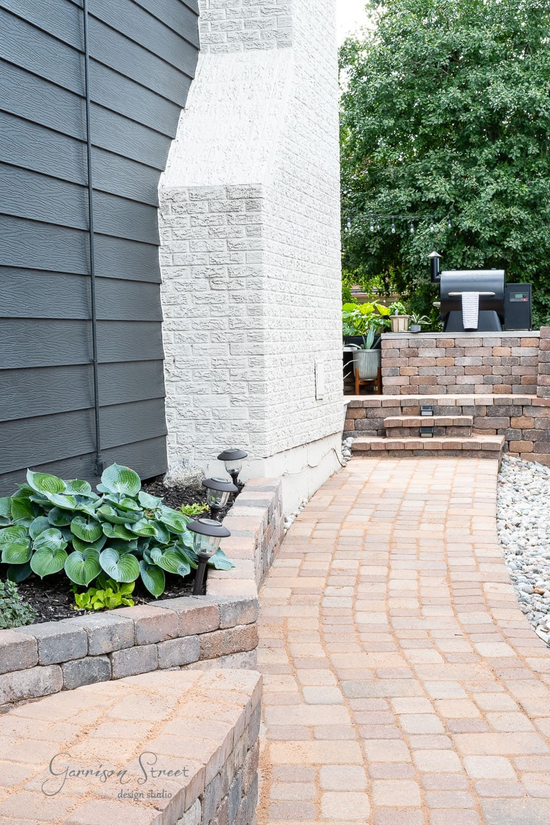 A landscaped backyard features a curved brick pathway bordered by neatly trimmed plants and small bushes. On the right, a DIY Traeger outdoor kitchen showcases a built-in grill on a paved raised area, with a background of trees and a brick retaining wall.