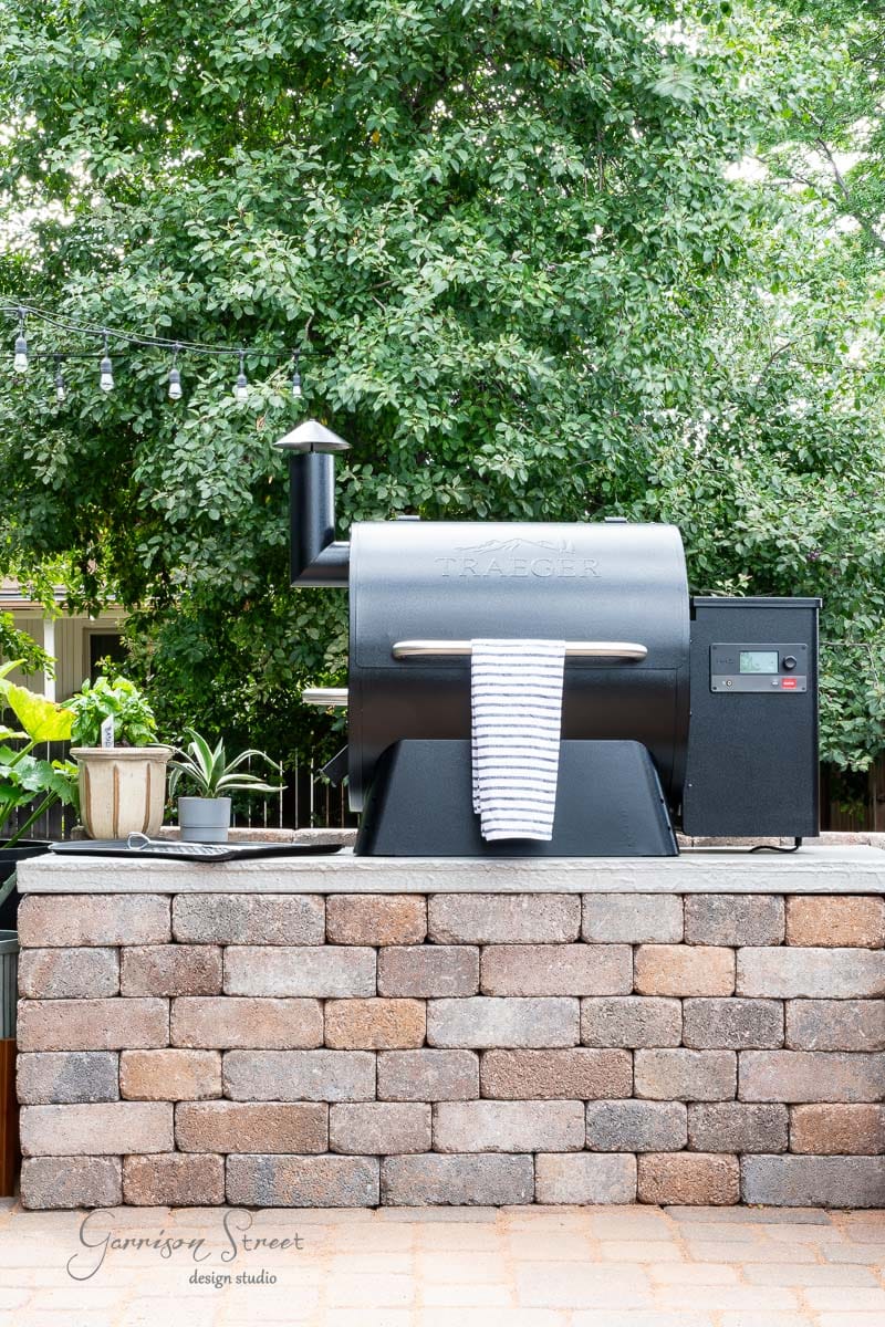 A black grill with a striped towel draped over its handle stands on a brick platform in this charming DIY outdoor kitchen. It's surrounded by lush green trees, and strung lights hang above, creating a cozy ambiance.
