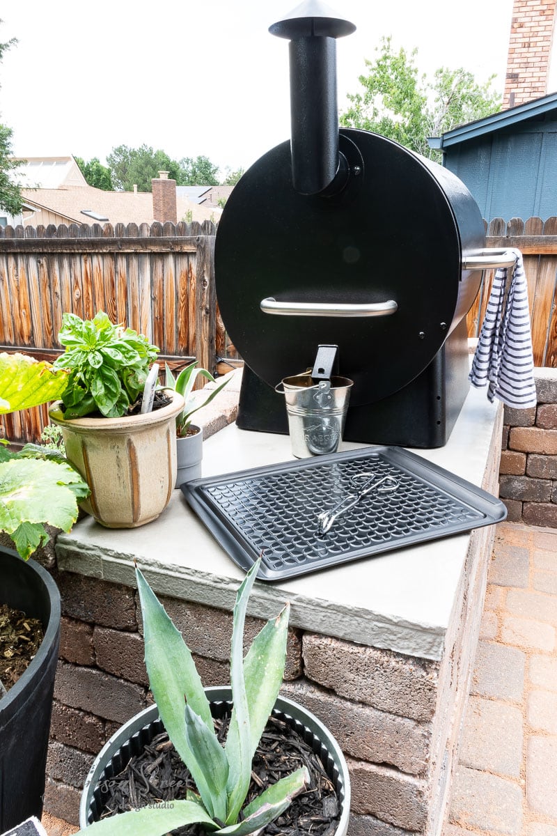 Outdoor grill with a bucket and tray on a brick platform, creating a DIY Traeger outdoor kitchen vibe. Potted plants, including agave and basil, surround the area. A towel hangs nearby on a fence, enclosing the cozy backyard setting.