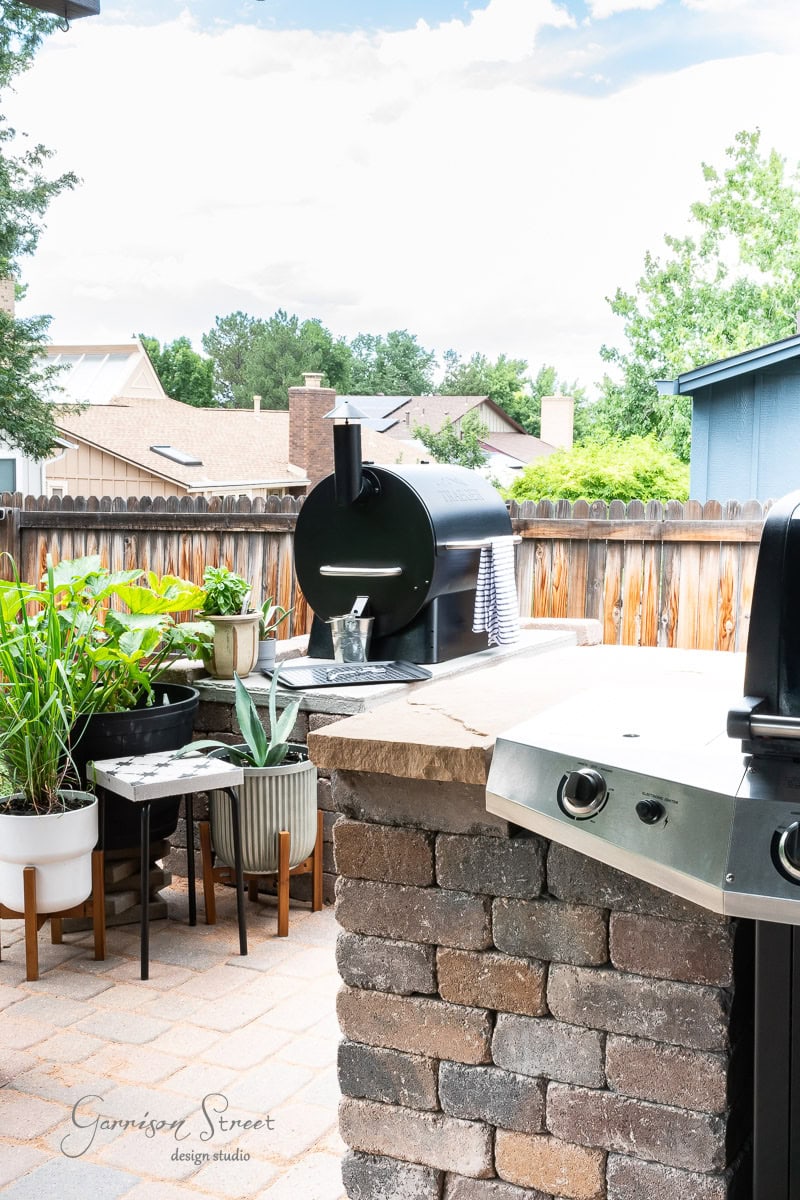 A backyard patio with a DIY outdoor kitchen featuring a grill, smoker, and Traeger setup. Potted plants sit on stone tables, and a wooden fence surrounds the area. Trees and rooftops are visible in the background under a cloudy sky.