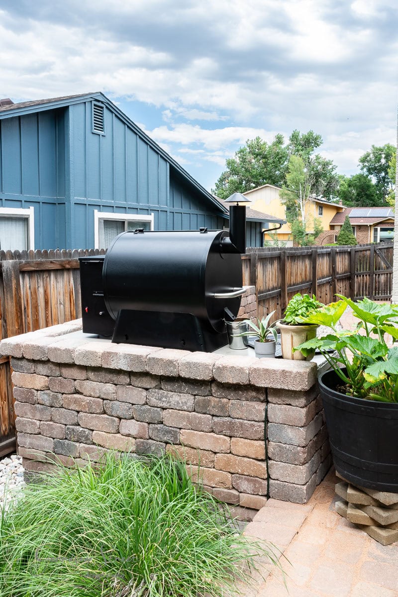 A black barbecue smoker sits on a stone wall in a DIY outdoor kitchen, surrounded by green plants in pots and a grassy patch. A blue house with a wooden fence is visible in the background, beneath a partly cloudy sky.