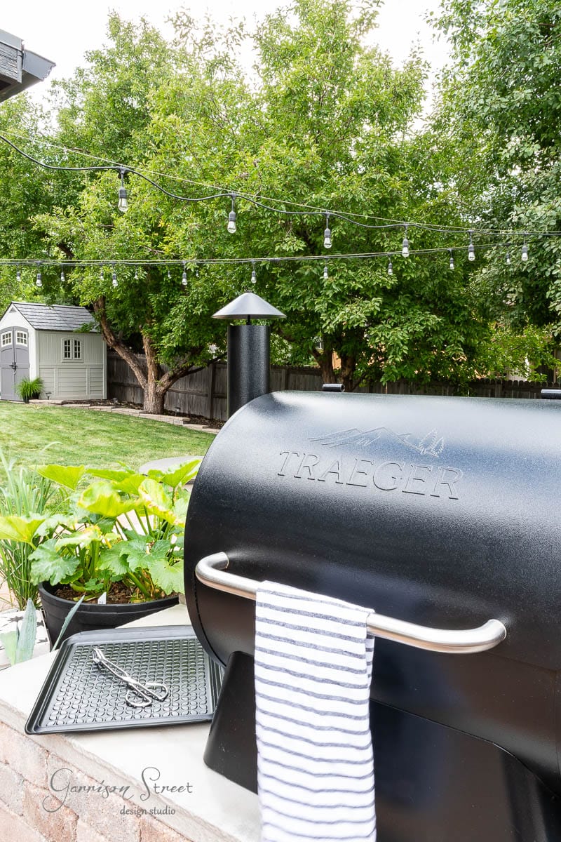A black Traeger grill with a striped towel draped over the handle is set in a DIY outdoor kitchen. In the background, lush green trees, a small garden shed, and string lights hanging across the yard create an inviting atmosphere. A tray with cooking tools is conveniently next to the grill.