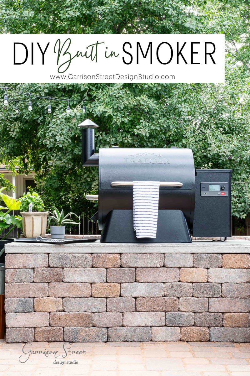 An outdoor built-in smoker sits on a brick platform, accented with a towel hanging on the front handle. Surrounded by lush greenery and twinkling string lights, it exudes charm. Above, a sign reads "DIY Traeger Outdoor Kitchen" with a website URL for those inspired to craft their own oasis.