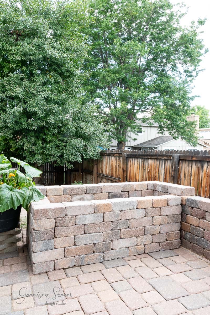 A raised garden bed made of stone bricks stands on a paved surface, perfectly complementing the DIY Traeger outdoor kitchen nearby. It is surrounded by a wooden fence, with lush green trees and plants under a clear sky.