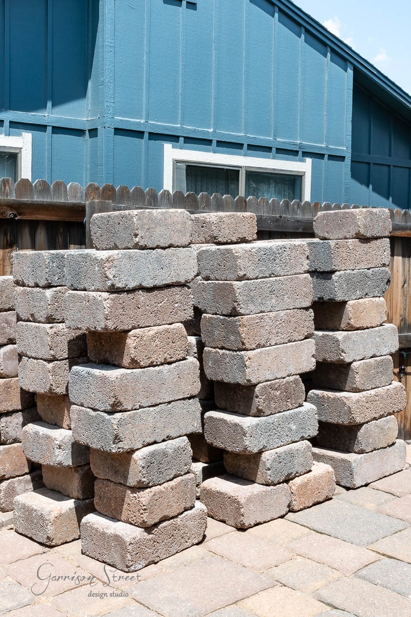 Stacks of rectangular concrete pavers are neatly arranged on a paved surface, ready for your next DIY project. A blue wooden building and a wooden fence frame the scene, perfect inspiration for creating an outdoor kitchen under the clear sky.