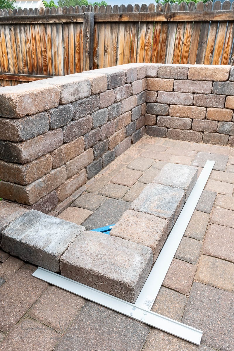 A DIY outdoor seating area is under construction, featuring a stone wall made of stacked bricks beside a Traeger outdoor kitchen. A level tool is laid across the top to ensure evenness. The bricks are positioned on a patio with a wooden fence in the background.