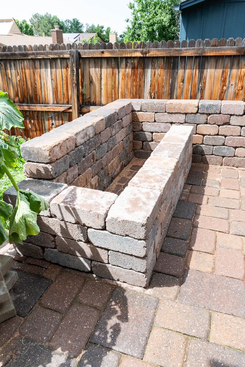 A DIY project unfolds with a raised garden bed under construction, featuring stacked stone bricks on a paved patio. Nearby, the Traeger outdoor kitchen hints at future culinary adventures. A wooden fence and greenery complete the rustic outdoor setting.