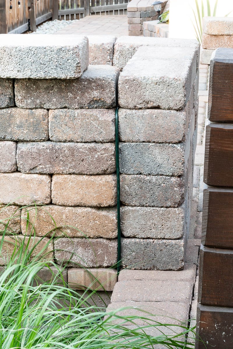 A stack of weathered gray bricks forms a corner in a garden, hinting at the beginnings of a DIY outdoor kitchen. Green leaves poke through the foreground, adding a touch of nature to the scene. The worn bricks suggest they've been in place for some time, ready to support culinary creations on your Traeger.