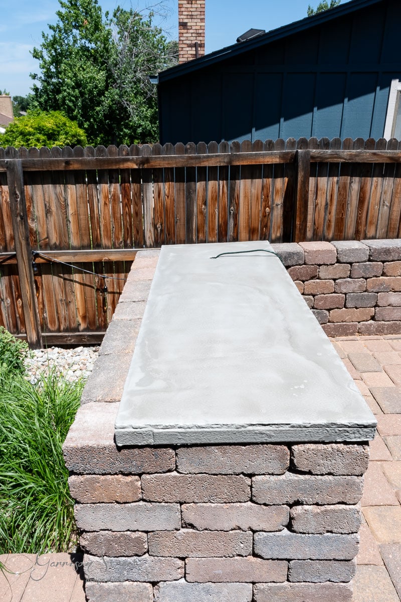 A DIY Traeger outdoor kitchen rests on a brick structure in a backyard with a wooden fence and lush greenery. A stone patio elegantly surrounds the area, while the blue sky highlights this inviting outdoor setting.
