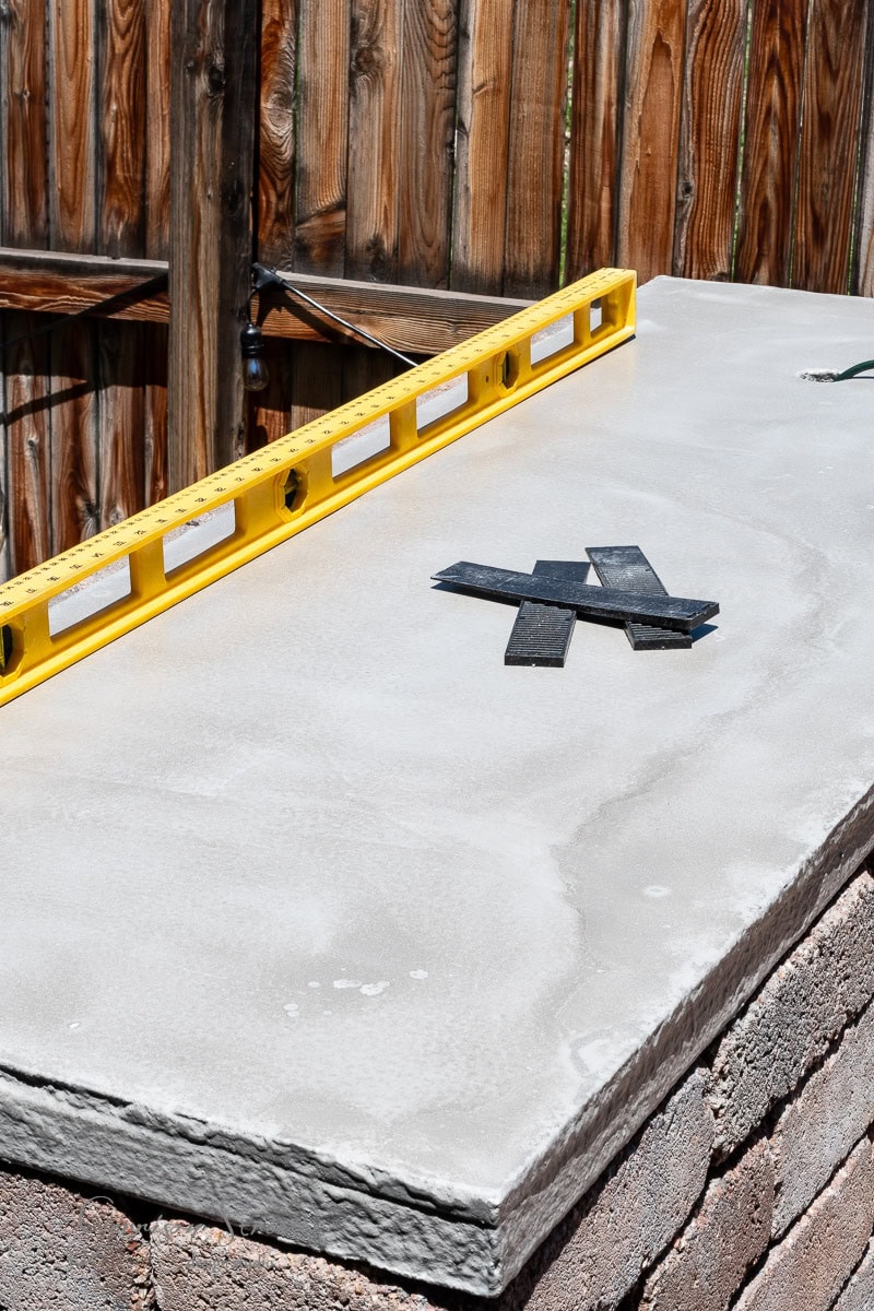 A freshly poured concrete slab sits atop a brick base, ready for your DIY Traeger outdoor kitchen. A yellow spirit level ensures precision, while two black metal X-shaped brackets rest on the smooth surface. A wooden fence completes this perfect setting for backyard culinary adventures.
