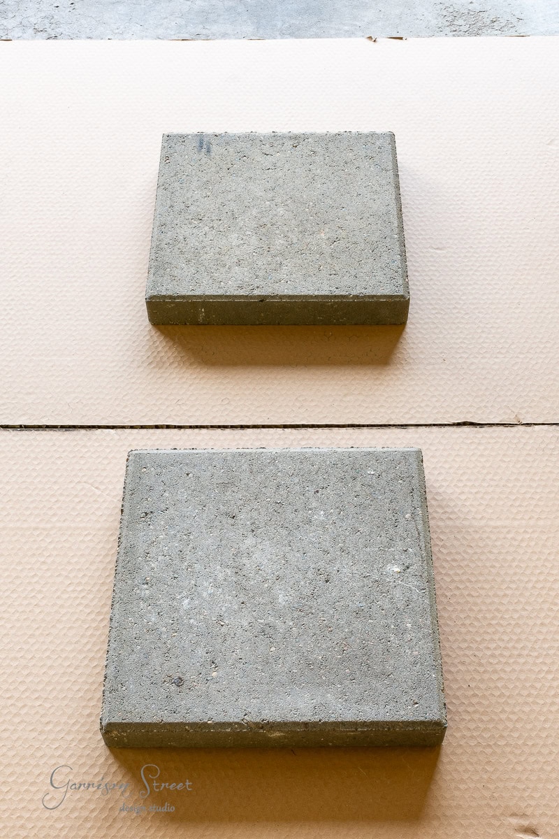 Two square concrete pavers rest on a textured, beige surface. They are aligned vertically with ample space between them, and the background reveals a subtle pattern.