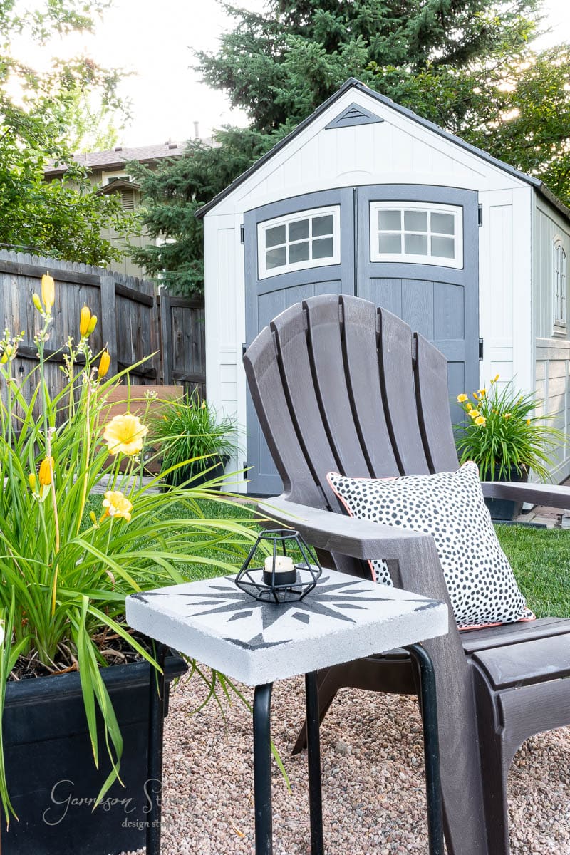 A backyard scene with a gray shed in the background is surrounded by lush green plants and bright yellow flowers. In the foreground, an Adirondack chair features a spotted cushion next to a DIY tile side table holding a candle holder.