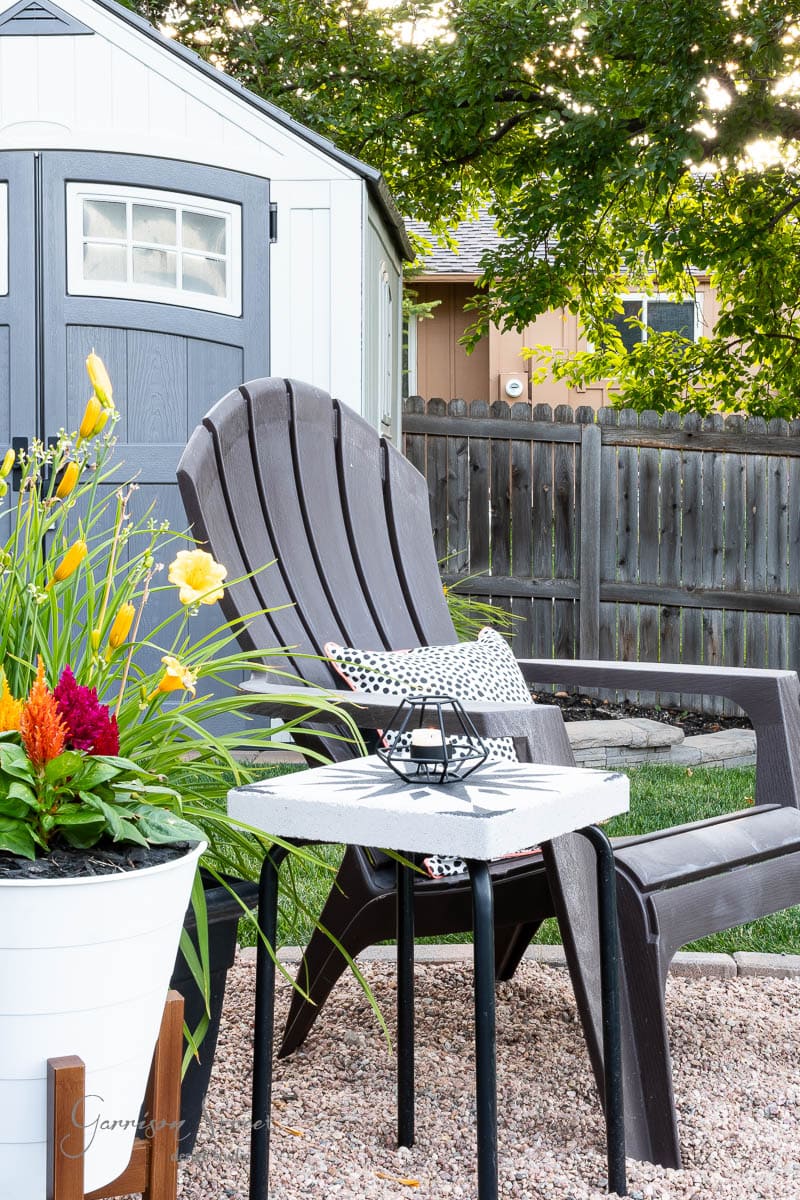 A backyard scene with a black Adirondack chair beside a DIY tile side table holding a candle. A white flower pot with colorful flowers sits on the gravel ground. In the background, there's a wooden fence, a tree, and a small shed.
