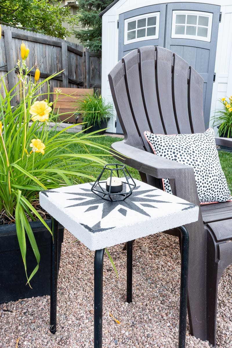 In a backyard setting, a black Adirondack chair is paired with a DIY tile side table showcasing a black geometric pattern. A small black candle holder rests on the table, complemented by yellow flowers and a white garden shed in the background.