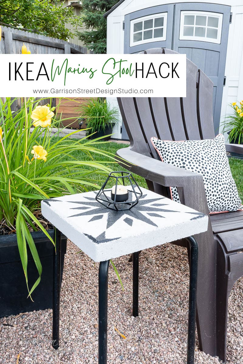 A small outdoor setup features a DIY IKEA stool transformed into a captivating tile side table with a sun motif. It's nestled on gravel alongside a black chair, a polka-dot pillow, vibrant yellow flowers, and a shed in the background. A geometric candle holder graces the tabletop.