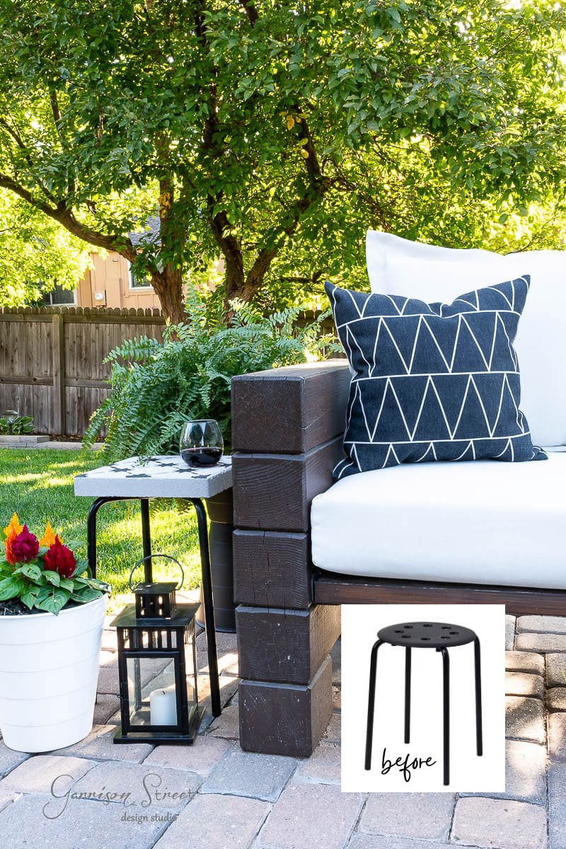 Outdoor patio with a cushioned bench featuring a geometric-patterned pillow. A small black stool labeled "before" is placed in the corner, paired with a DIY tile side table. A potted plant, lantern, wine glass, and greenery surround the area. Sunlight filters through the trees.