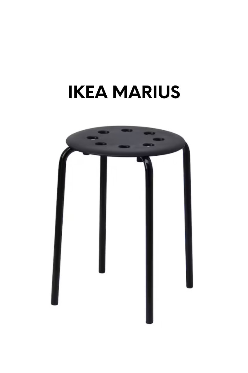 A black IKEA MARIUS stool with a round seat featuring circular cutouts and four slender metal legs stands against a plain white background.