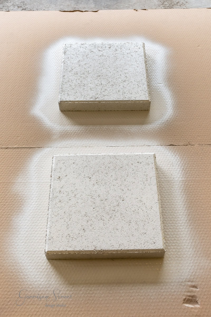 Two square stone tiles are artfully arranged on a textured surface, surrounded by a light spray of white paint forming circular halos. The backdrop is cardboard.