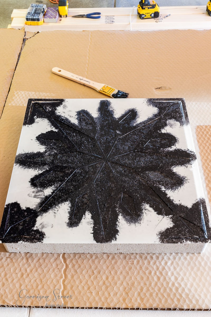 A square resin block with a black starburst pattern sits on a cardboard surface. A paintbrush with black bristles lies nearby, while construction tools are visible in the background.