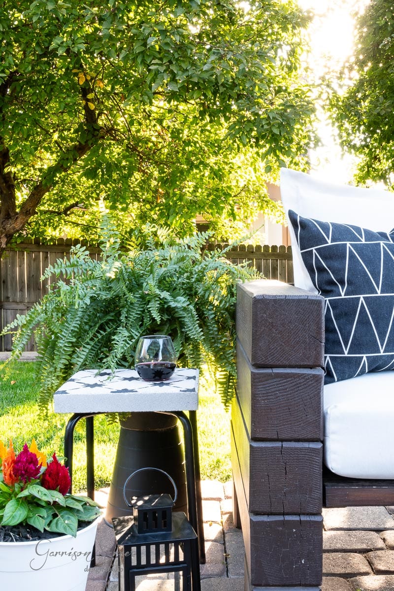 A cozy outdoor patio scene features a cushioned sofa with a geometric black and white pillow, a lush fern on a DIY tile side table, a glass of wine, and a black lantern on the brick floor. Sunlight gently filters through the trees in the background.