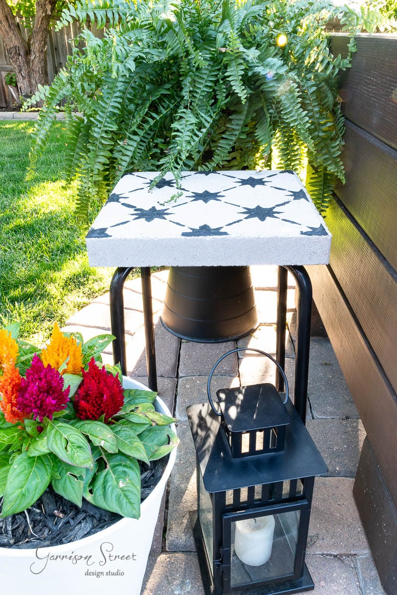 A small DIY tile side table with a black and white geometric patterned top stands on a brick patio. Nearby is a potted plant with colorful flowers and a black lantern. A large fern in the background adds greenery to the scene.
