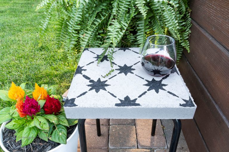 A small outdoor tile side table with a star-patterned white surface holds a glass of red wine. It is surrounded by a lush green fern and a colorful flower pot with vibrant red, orange, and yellow blossoms on a brick patio.
