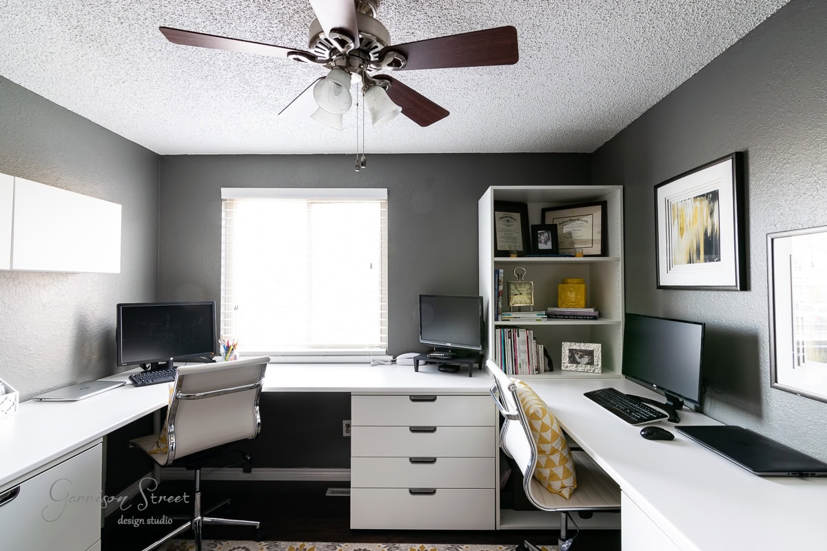 A modern DIY home office for two features dark gray walls and a textured ceiling with two white L-shaped desks, chairs, and dual computer monitors. The room is lit by a ceiling fan and a window with blinds. Shelves display books and decor items, creating an inspiring workspace.