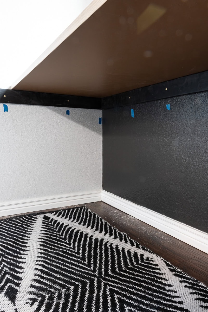 A corner of a DIY home office for two showcases two-tone painted walls—one white, the other dark gray. An abstract black and white rug graces the floor, while blue tape pieces add a creative touch above the baseboards on the dark wall.