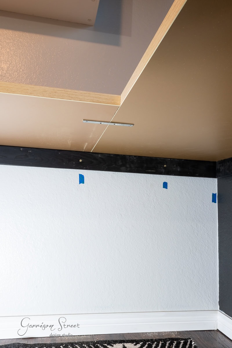 A ceiling with recessed lighting and a metal bracket is shown, suggesting a DIY home office for two. The wall is white with the bottom partially painted black. Pieces of blue painter's tape are placed on the wall, and a black and white area rug completes the setup at the bottom.