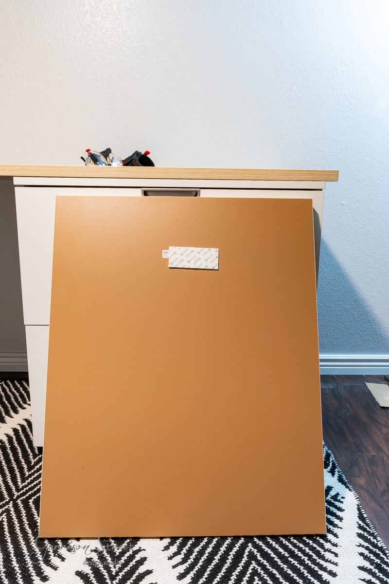 A brown wooden board leans against a white desk with a light wooden top, crafting the perfect DIY home office setup. On the desk are some tools, while the floor boasts a black and white zigzag patterned rug. The backdrop is completed by a crisp white wall.